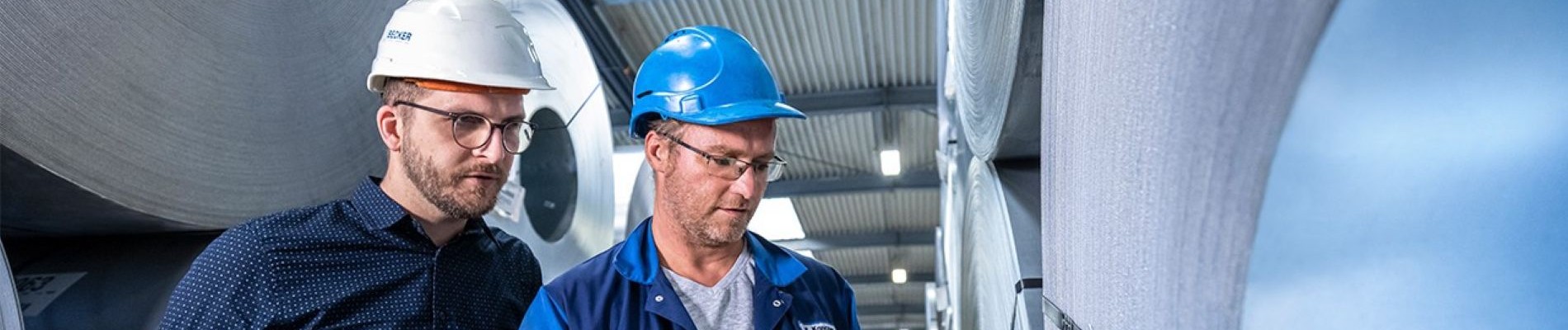 Zwei Arbeiter mit Schutzhelmen inspizieren Maschinen in einer industriellen Umgebung und konzentrieren sich dabei auf Details der Ausrüstung.