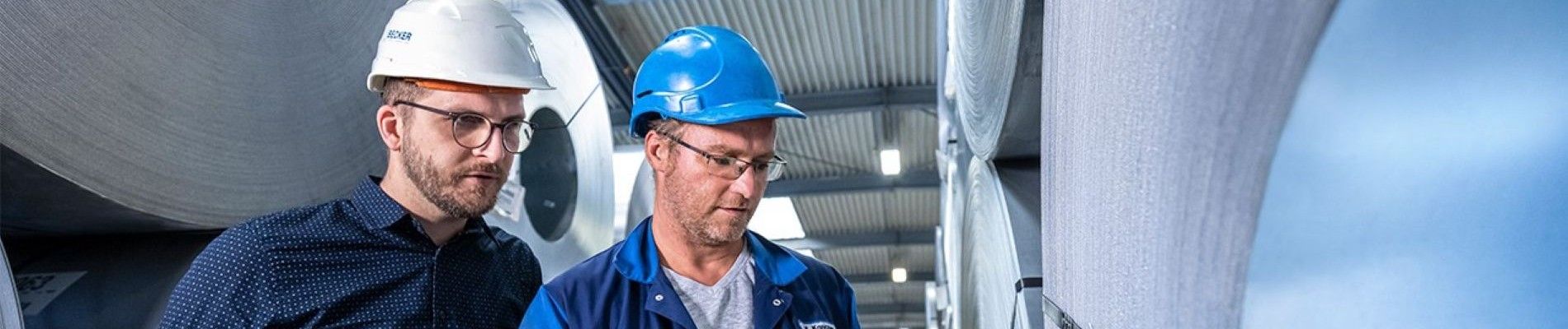 Zwei Arbeiter mit Schutzhelmen inspizieren Maschinen in einer industriellen Umgebung und konzentrieren sich dabei auf Details der Ausrüstung.