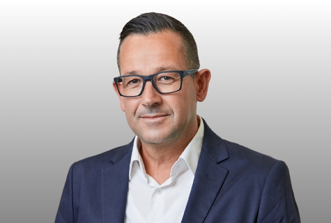 Man with short dark hair and glasses wearing a navy suit and white shirt, smiling slightly against a light gray background.