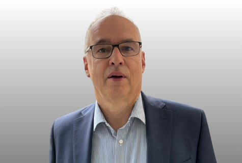 Man with glasses wearing a blue suit and striped shirt, standing against a gradient gray background.