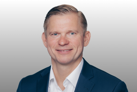 Man in a blue suit and white shirt smiling, with short blond hair, against a gray background.
