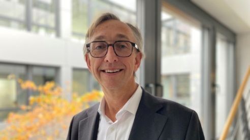 Eine lächelnde Person mit Brille und Anzug steht in einem Innenraum in der Nähe großer Fenster, durch die man das Herbstlaub draußen sehen kann.