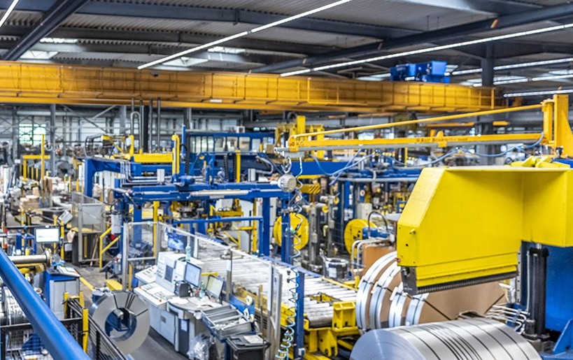 Innenraum einer Industrieanlage mit leuchtend gelben und blauen Maschinen, Coils und Laufkränen unter einer hohen Decke.